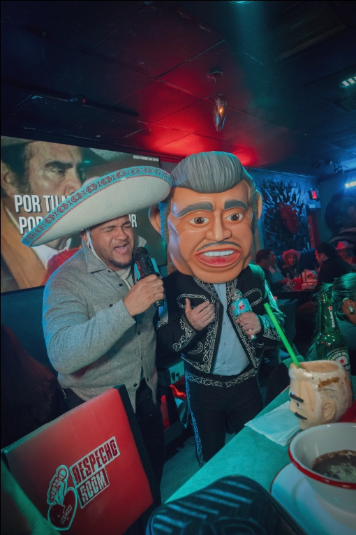 A customer singing karaoke with a sombrero next to a mascot with a giant head in the lively atmosphere of Despecho Room bar in Garfield, NJ.
