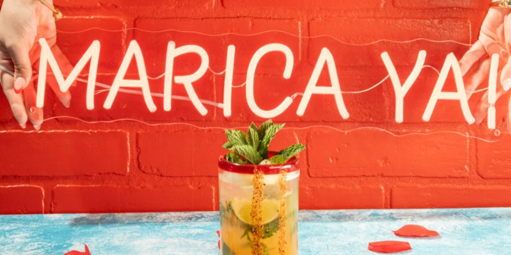 A mojito-style cocktail in front of a neon sign that reads 'Marica Ya!', a popular Colombian slang phrase used at the Despecho Room bar in Garfield, NJ.